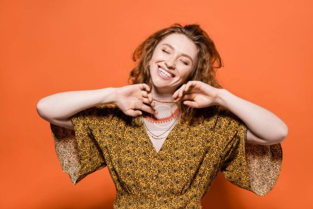 Positive young red haired model in yellow blouse with abstract pattern touching necklaces and standing with closed eyes on orange background, stylish casual outfit and summer vibes concept, Youth Culture