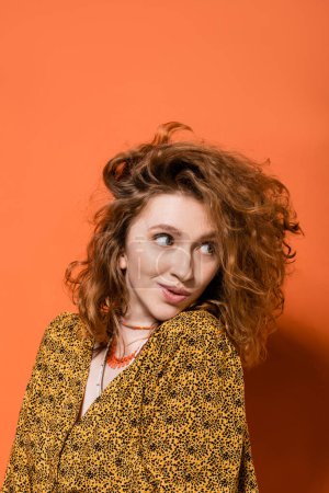 Astonished young red haired woman in yellow blouse with abstract pattern and necklaces looking away and standing on orange background, stylish casual outfit and summer vibes concept, Youth Culture