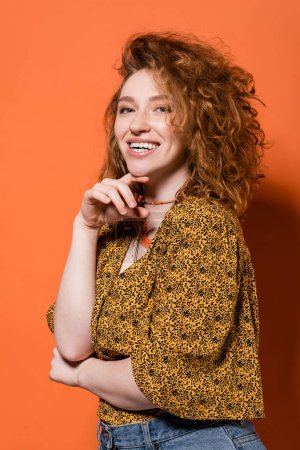 Photo for Happy young red haired woman in necklaces, yellow blouse with abstract pattern looking at camera and posing on orange background, stylish casual outfit and summer vibes concept, Youth Culture - Royalty Free Image