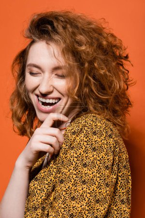 Portrait de jeune femme rousse positive en chemisier jaune avec motif abstrait debout les yeux fermés sur fond orange, tenue décontractée élégante et concept d'ambiance estivale, Culture jeunesse