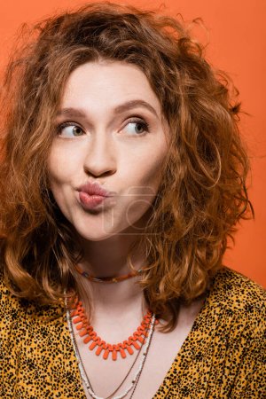 Portrait of young red haired woman in yellow blouse and necklaces pouting lips and looking away while standing isolated on orange, stylish casual outfit and summer vibes concept, Youth Culture