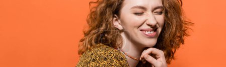 Gefallen junge rothaarige Frau mit natürlichem Make-up berühren Halskette und posiert in Bluse mit abstrakten Muster isoliert auf orange, stilvolle lässige Outfit und Sommer-Atmosphäre Konzept, Banner