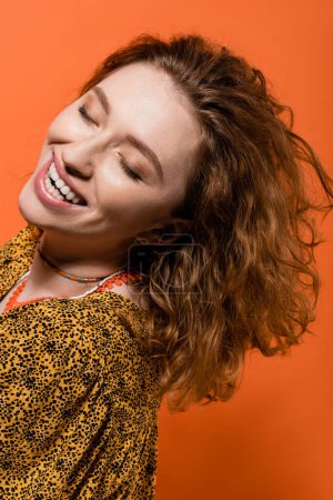 Porträt einer glücklichen jungen rothaarigen Frau in Halsketten und trendiger Bluse mit abstraktem Muster, die isoliert auf orangefarbenem, stylischem Casual-Outfit und sommerlichem Vibes-Konzept steht, Jugendkultur