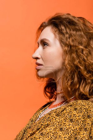 Photo for Side view of stylish redhead woman in necklaces and yellow blouse with abstract pattern looking away while standing isolated on orange, stylish casual outfit and summer vibes concept, Youth Culture - Royalty Free Image