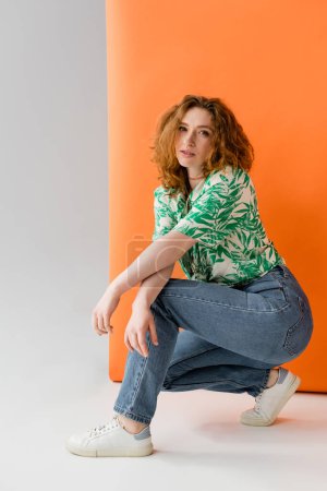 Full length of stylish redhead woman in blouse with floral pattern and jeans looking at camera while posing on orange and grey background, trendy casual summer outfit concept, Youth Culture