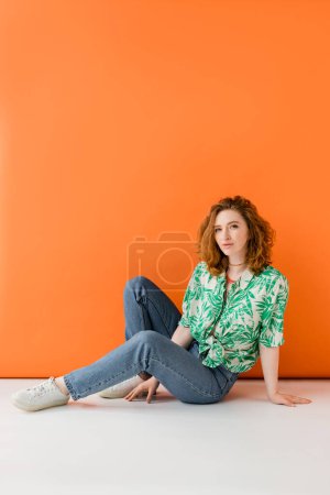 Volle Länge der jungen rothaarigen Frau mit natürlichem Make-up posiert in Bluse mit Blumenmuster und Jeans, während sie auf orangefarbenem Hintergrund sitzt, trendiges lässiges Sommer-Outfit-Konzept, Jugendkultur