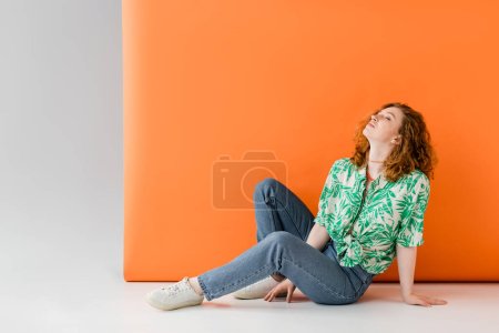 Mujer de pelo rojo joven y relajada con los ojos cerrados en blusa con estampado floral y jeans sentados sobre fondo gris y naranja, concepto de atuendo de verano casual de moda, cultura juvenil