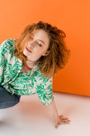 Stylish young redhead woman in blouse with floral pattern, necklaces and jeans posing with closed eyes on orange background, trendy casual summer outfit concept, Youth Culture