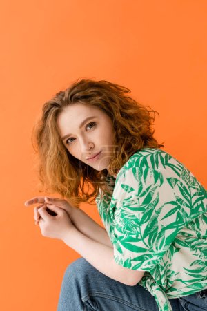 Portrait of stylish and confident redhead model in blouse with floral pattern and jeans looking at camera isolated on orange, trendy casual summer outfit concept, Youth Culture