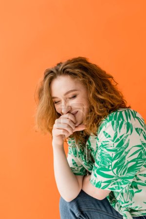 Fröhliche junge rothaarige Frau mit geschlossenen Augen in Bluse mit Blumenmuster und Jeans lacht, während sie isoliert auf orangefarbenem, trendigem Sommeroutfit-Konzept posiert, Jugendkultur