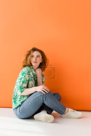 Longitud completa de mujer pelirroja joven en blusa elegante con patrón floral y jeans mirando hacia otro lado mientras está sentado sobre fondo naranja, concepto de traje de verano casual de moda, Cultura Juvenil