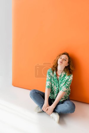 Femme aux cheveux rouges décontractés en jeans, chemisier moderne avec motif floral et colliers assis sur fond gris et orange, concept de tenue estivale décontractée tendance, Culture jeunesse