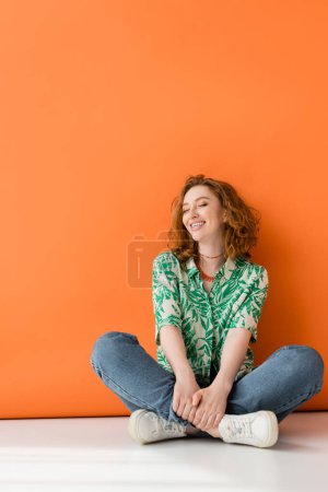 Volle Länge der positiven jungen rothaarigen Frau in Bluse mit Blumenmuster und Jeans sitzend mit geschlossenen Augen auf orangefarbenem Hintergrund, trendiges lässiges Sommeroutfit-Konzept, Jugendkultur
