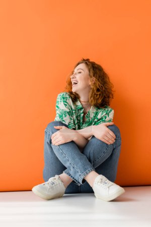 Fröhliches junges rothaariges Model in stylischer Bluse mit Blumenmuster und moderner Jeans, die die Knie berührt und auf orangefarbenem Hintergrund sitzt, trendiges lässiges Sommeroutfit-Konzept, Jugendkultur