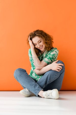 Volle Länge der stilvollen jungen rothaarigen Frau in Bluse mit Blumenmuster und Jeans lächelnd auf orangefarbenem Hintergrund, trendiges lässiges Sommeroutfit-Konzept, Jugendkultur