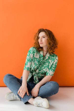 Mujer pelirroja joven confiada en blusa moderna con patrón floral y jeans mirando hacia otro lado mientras está sentada sobre fondo naranja, concepto de traje de verano casual de moda, Cultura Juvenil