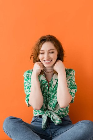 Fröhliches junges rothaariges Model in Jeans und Bluse mit floralem Muster, das Halsketten berührt, während es auf orangefarbenem Hintergrund sitzt, trendiges lässiges Sommer-Outfit-Konzept, Jugendkultur