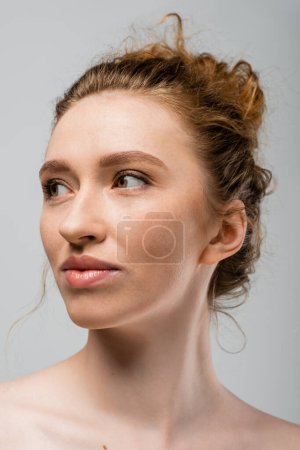 Porträt einer jungen rothaarigen und sommersprossigen Frau mit natürlichem Make-up und nackten Schultern, die wegschaut und isoliert auf einem grauen, natürlichen Schönheits- und Individualitätskonzept steht, Jugendkultur