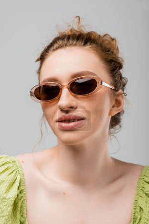 Stylische junge rothaarige und sommersprossige Frau in moderner und lässiger grüner Bluse und Sonnenbrille, lächelnd und isoliert auf grauem, trendigem Sonnenschutzkonzept stehend, Jugendkultur