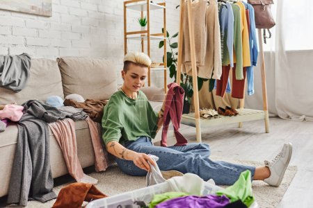 tattooed woman sorting wardrobe items while sitting on floor in living room near couch and rack with clothes, decluttering and reducing, sustainable living and mindful consumerism concept