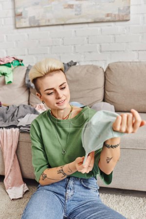 wardrobe items decluttering, young and cheerful woman with tattoo and trendy hairstyle holding cap near couch with clothes in living room, sustainable living and mindful consumerism concept