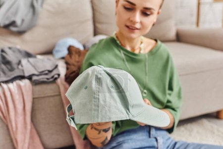 sorting clothes, young woman holding stylish cap during wardrobe items decluttering near couch at home, blurred background, sustainable living and mindful consumerism concept