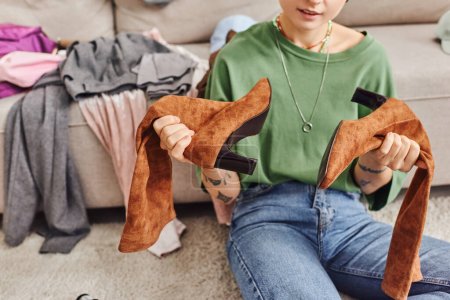Téléchargez les photos : Vue partielle de la jeune femme tatouée en vêtements décontractés assis sur le sol et tenant des bottes en daim, des vêtements de tri, une garde-robe débordante à la maison, une vie durable et un concept de consumérisme conscient - en image libre de droit