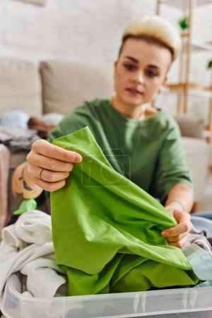junge tätowierte Frau reduziert Kleiderschrank, sortiert Kleidung und hält grüne Kleidungsstücke zu Hause im Wohnzimmer, verschwommener Hintergrund, nachhaltiges Leben und bewusstes Konsumkonzept