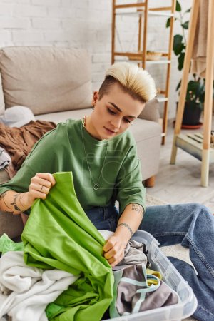 young woman with tattoo and trendy hairstyle holding green garment near plastic container while sorting clothes and reducing wardrobe at home, sustainable living and mindful consumerism concept