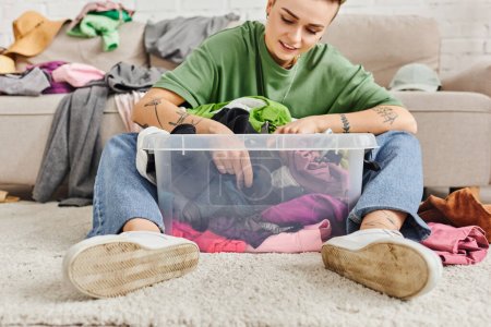 smiling and tattooed woman sitting on floor near plastic container with thrift store finds, decluttering process, clothes sorting, sustainable living and mindful consumerism concept