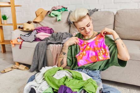 mujer sonriente sosteniendo la parte superior colorida mientras decluttering artículos de armario cerca del sofá con ropa en la sala de estar moderna, peinado de moda, tatuaje, vida sostenible y concepto de consumismo consciente