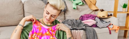 smiling woman with trendy hairstyle and tattoo looking at colorful top near couch in living room, sorting clothes, decluttering process, sustainable living and mindful consumerism concept, banner