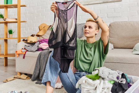 stylish and tattooed woman looking at bodysuit, sitting on floor near couch with wardrobe items, sorting clothes, decluttering process, sustainable living and mindful consumerism concept