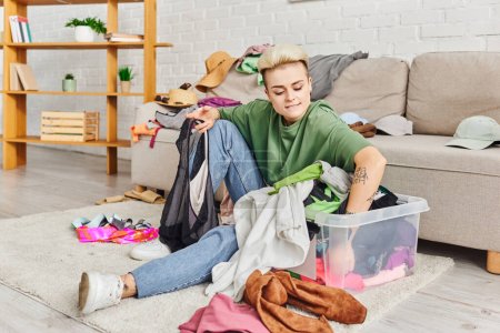 young woman sorting clothes in plastic container while sitting on floor near couch in living room, trendy hairstyle, tattoo, decluttering process, sustainable living and mindful consumerism concept