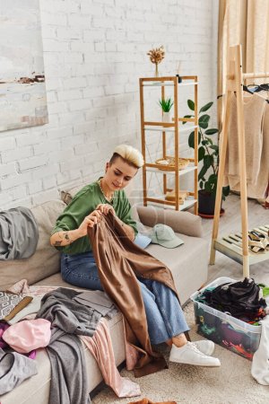 young tattooed woman holding leather pants while sitting on couch near clothes, racks and plants in modern living room, decluttering process, sustainable living and mindful consumerism concept
