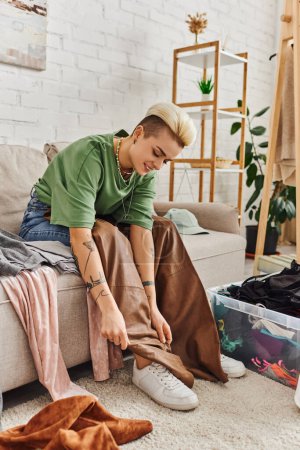 positive tattooed woman sitting on couch, looking at leather pants, decluttering clothes near plastic container, racks and green plants at home, sustainable living and mindful consumerism concept