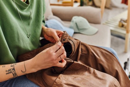 vista recortada de la joven mujer tatuada abotonar pantalones de cuero mientras está sentado en el sofá cerca de artículos de armario durante el proceso de decluttering, vida sostenible y concepto de consumismo consciente
