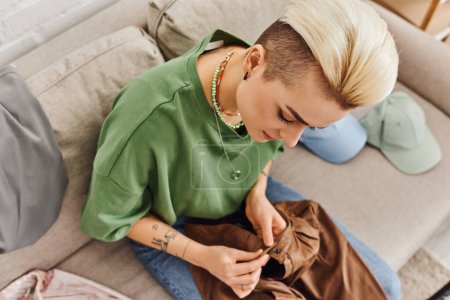 vista superior de la mujer elegante con peinado de moda y tatuaje sentado en el sofá cerca de la ropa y abotonar pantalones de cuero, proceso de decluttering, vida sostenible y concepto de consumismo consciente