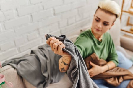 Téléchargez les photos : Jeune femme tatouée assise sur le canapé dans le salon et tenant un pantalon gris tout en réduisant les articles de garde-robe à la maison, coiffure à la mode, tatouage, vie durable et concept de consumérisme conscient - en image libre de droit