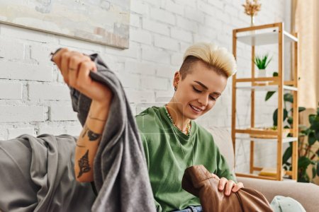 carefree woman with tattoo and trendy hairstyle sorting pre-loved items and second-hand clothing in modern living room, wardrobe reducing, sustainable living and mindful consumerism concept