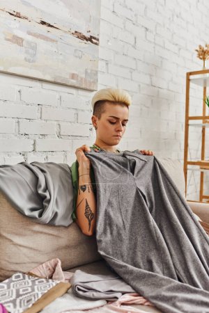 Foto de Mujer joven reflexiva con peinado de moda y tatuaje sentado en el sofá cerca de artículos de armario y la celebración de pantalones grises, clasificación de ropa, decluttering, vida sostenible y concepto de consumismo consciente - Imagen libre de derechos