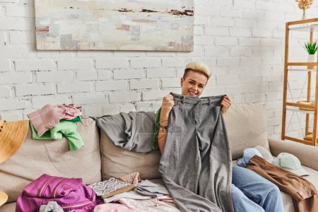 overjoyed tattooed woman holding grey pants and looking at camera near clothes on couch in living room, sorting wardrobe items, home decluttering, sustainable living and mindful consumerism concept