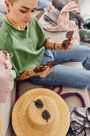 Photo for High angle view of stylish and tattooed woman holding sunglasses near straw hat and wardrobe items on couch at home, sorting clothes, sustainable living and mindful consumerism concept - Royalty Free Image