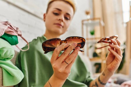 low angle view of young tattooed woman holding trendy sunglasses while sorting and decluttering clothing and pre-loved items, blurred background, sustainable living and mindful consumerism concept