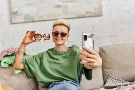 Téléchargez les photos : Femme tatouée heureux assis sur le canapé près des articles de garde-robe et de prendre selfie avec des lunettes de soleil sur smartphone pour l'échange en ligne, la vie durable et le concept de consumérisme conscient - en image libre de droit