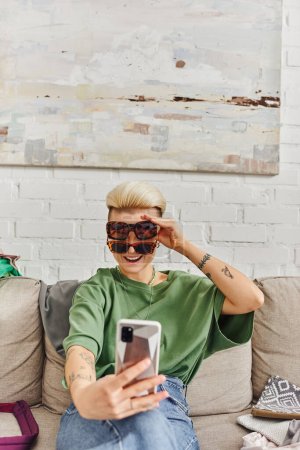 young, excited and tattooed woman taking selfie in stylish sunglasses on smartphone near clothes on couch for online swap in social media, sustainable living and mindful consumerism concept