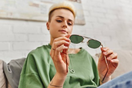 Low-Winkelansicht einer jungen tätowierten Frau mit trendiger Frisur, die zu Hause eine modische Sonnenbrille anschaut, um ihr Lieblingsobjekt, nachhaltiges Leben und bewusstes Konsumkonzept zu erklären