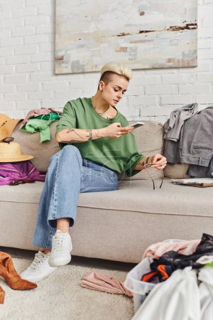 online swap on virtual marketplace, tattooed woman in casual clothes taking photo of sunglasses on couch near wardrobe items in modern living room, sustainable living and mindful consumerism concept