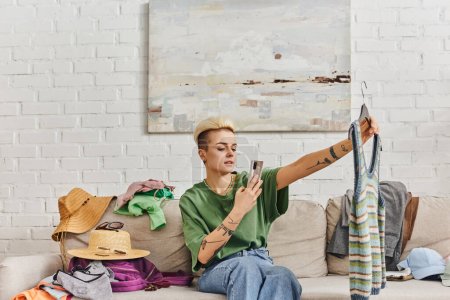 Tätowierte Frau mit Smartphone fotografiert gestrickte Tanktop auf Couch in der Nähe von Strohhüten und Kleidung, Online-Austausch, Kreislaufwirtschaft, nachhaltiges Leben und achtsames Konsumkonzept