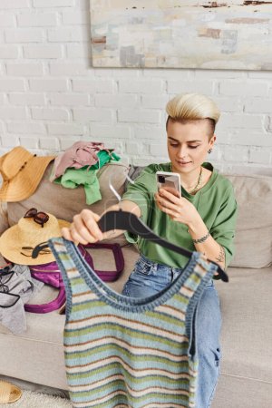 Online-Kleiderbörse, positiv tätowierte Frau mit Handy auf gestricktem Tanktop neben Strohhüten und Kleidung auf Couch, nachhaltiges Leben und bewusstes Konsumkonzept